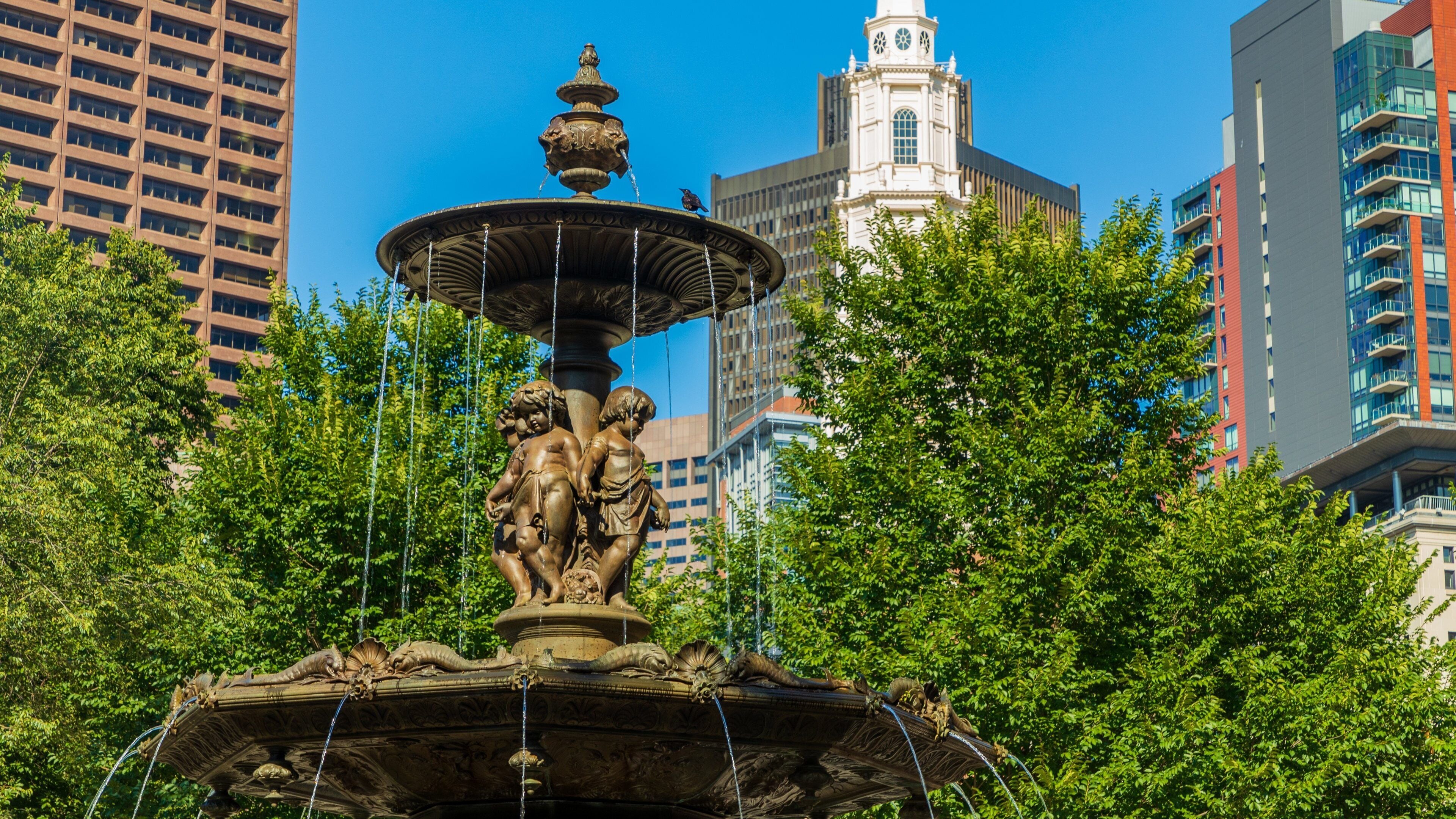 Uma fonte com estátuas num parque, com edifícios altos ao fundo.