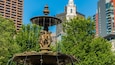A fountain with statues in a park with tall buildings in the background.