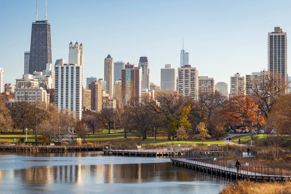 A city skyline with modern skyscrapers and a park with autumn trees and a lake.