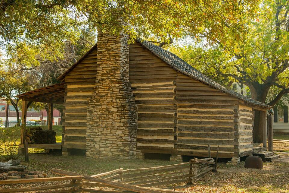 A rustic log cabin with a stone chimney, surrounded by trees and a wooden fence.
