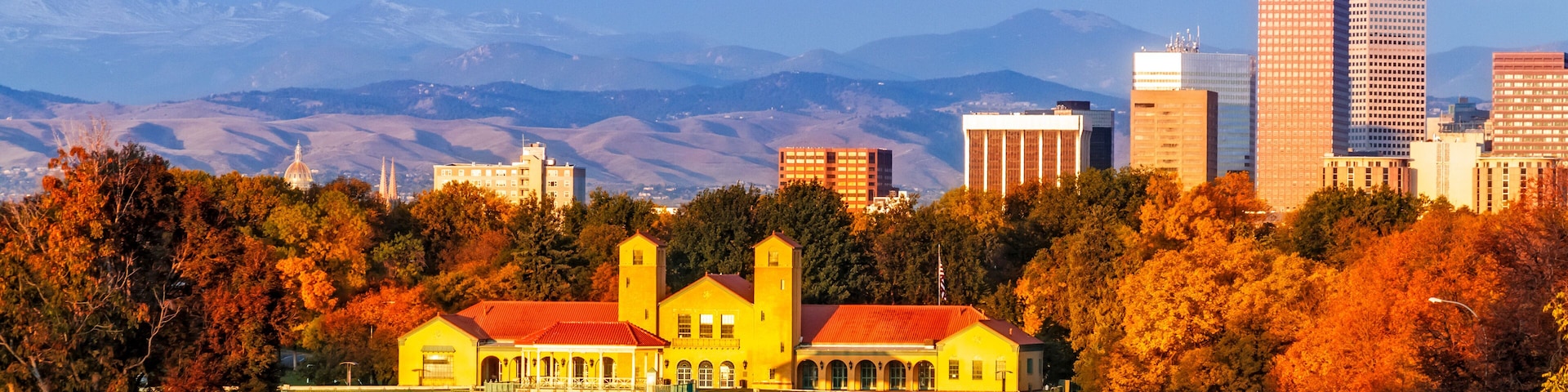 A cityscape with a prominent yellow building reflecting in a lake, surrounded by autumn trees and mountains in the background.
