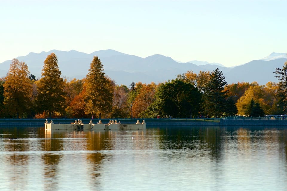 A calm lake with ducks, surrounded by autumn-colored trees and mountains in the background.