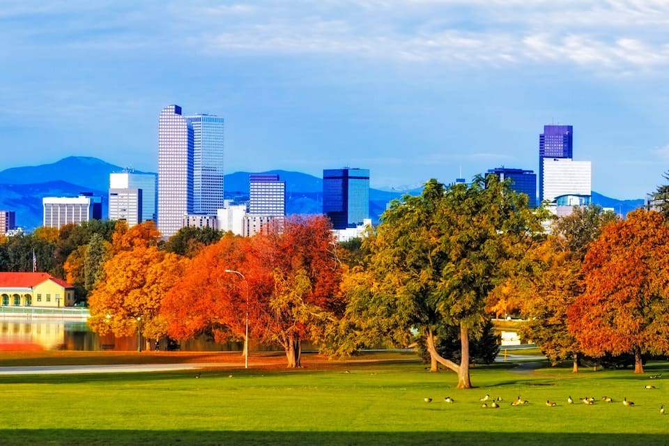 A park with autumn trees, a lake, and a city skyline with mountains in the background.