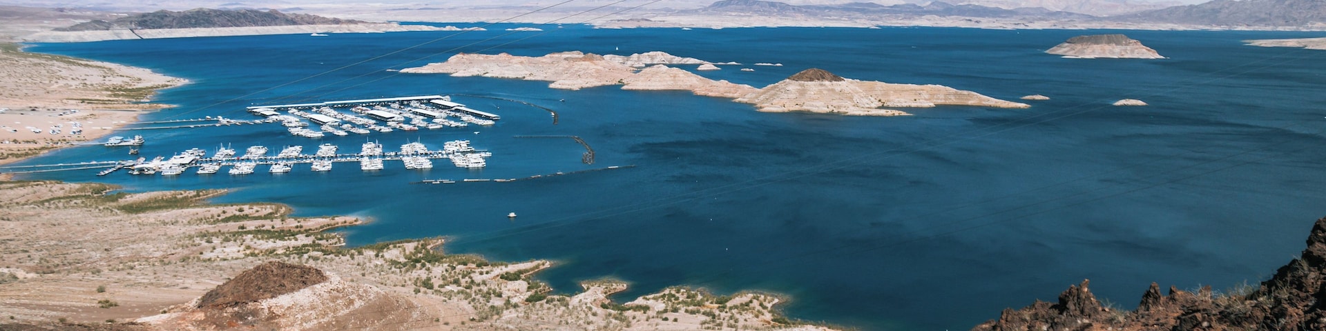 A coastal marina with numerous boats docked, surrounded by rocky terrain and distant mountains.