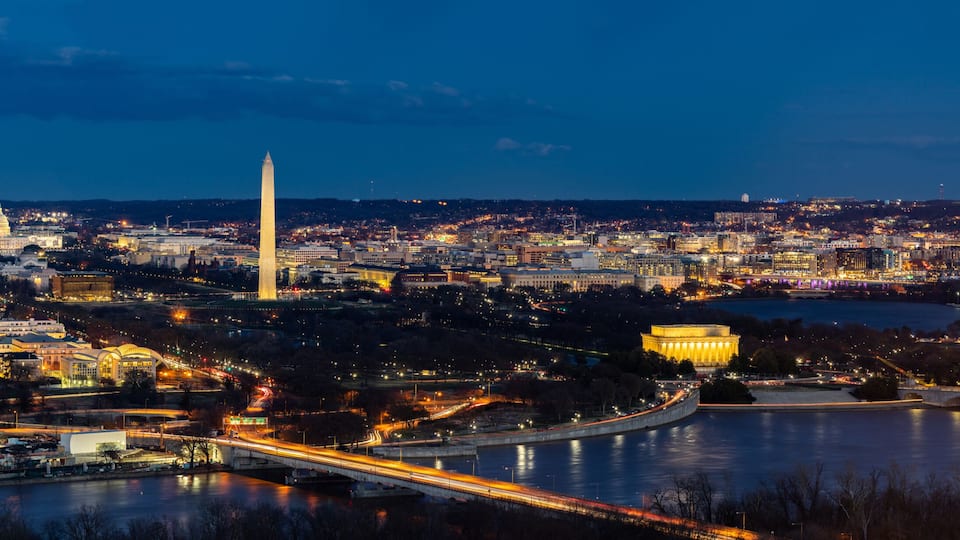 A nighttime cityscape with prominent landmarks including a tall obelisk, a large domed building, and a historic monument.