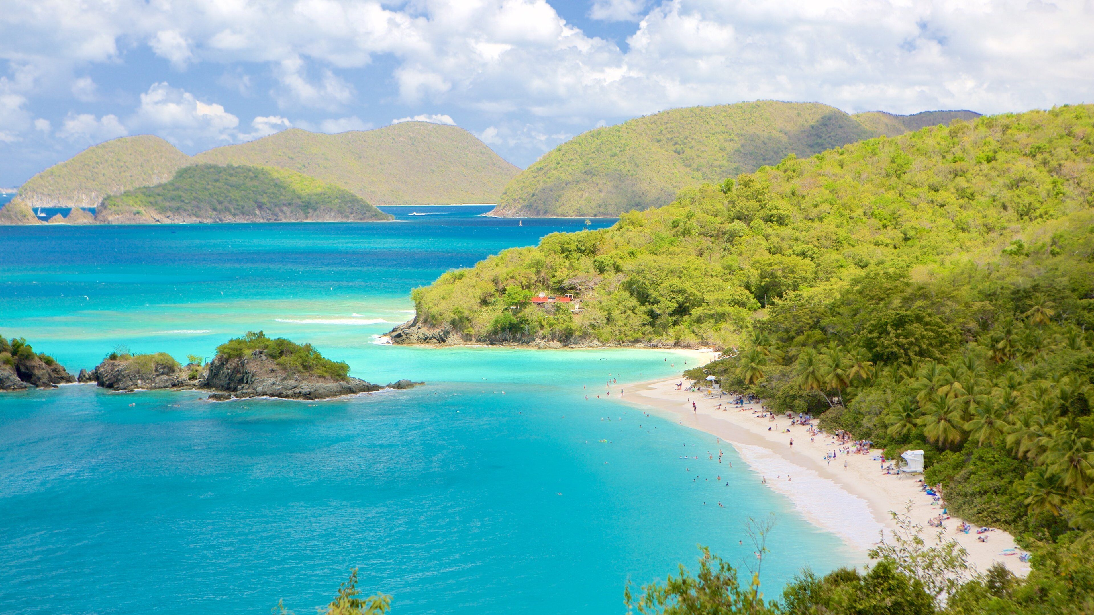 A tropical beach with clear turquoise waters, a sandy shore, and lush green hills in the background.