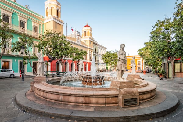 Une fontaine ornée de statues, entourée de bâtiments historiques aux façades rouges et jaunes.