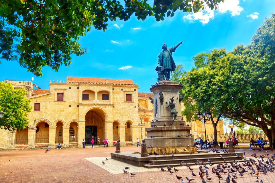 A historic plaza with a statue, a building with arches, and pigeons.