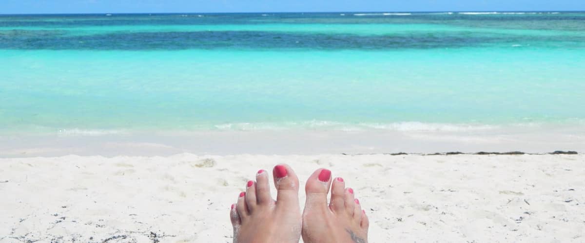 A pair of feet with red-painted toenails on a sandy beach with turquoise water and a clear blue sky.