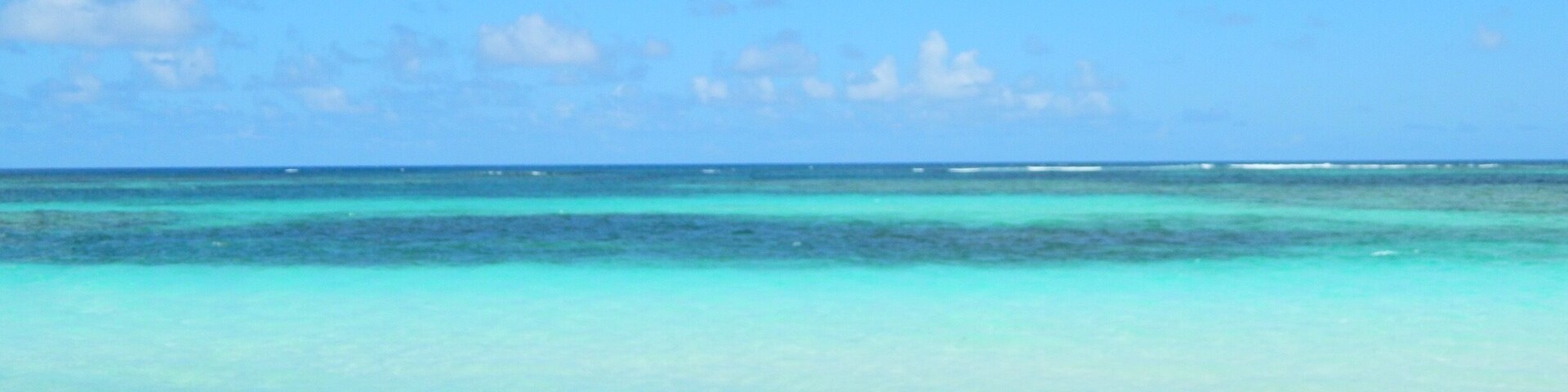 A pair of feet with red-painted toenails on a sandy beach with turquoise water and a clear blue sky.