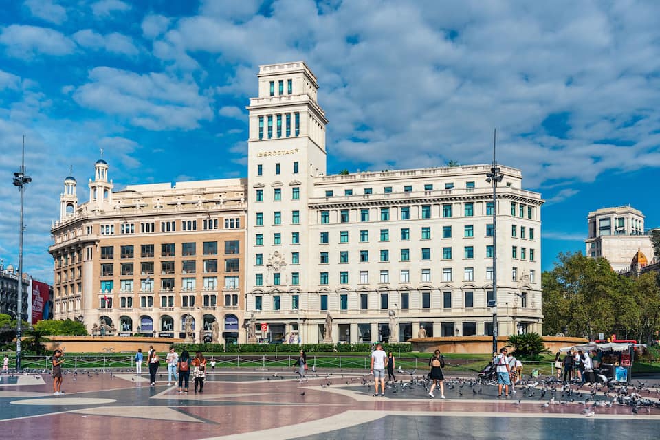 Un grand bâtiment historique à étages, surmonté d’une tour imposante, entouré d’une place publique où se promènent des gens et où volent des pigeons.