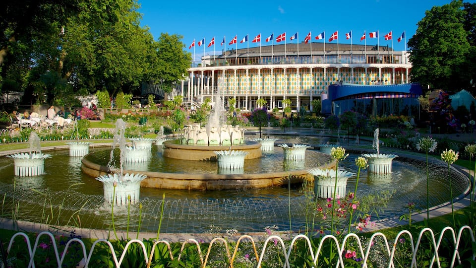 A large fountain with multiple tiers, surrounded by a garden with various flowers and plants. In the background, there is a building with multiple flags and a colorful facade.