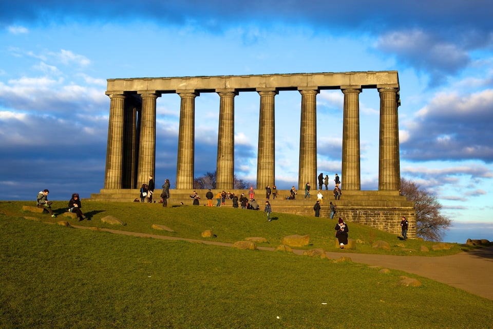 Um grande edifício clássico com colunas imponentes, cercado por uma área gramada onde pessoas caminham e se sentam.