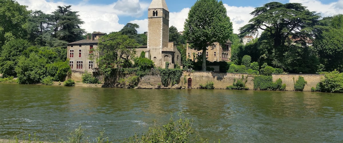 A historic stone tower by a river, surrounded by lush greenery and traditional buildings.