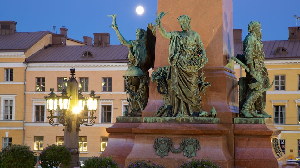 A bronze statue of a woman holding a torch, flanked by two smaller statues, with a building and a street lamp in the background.