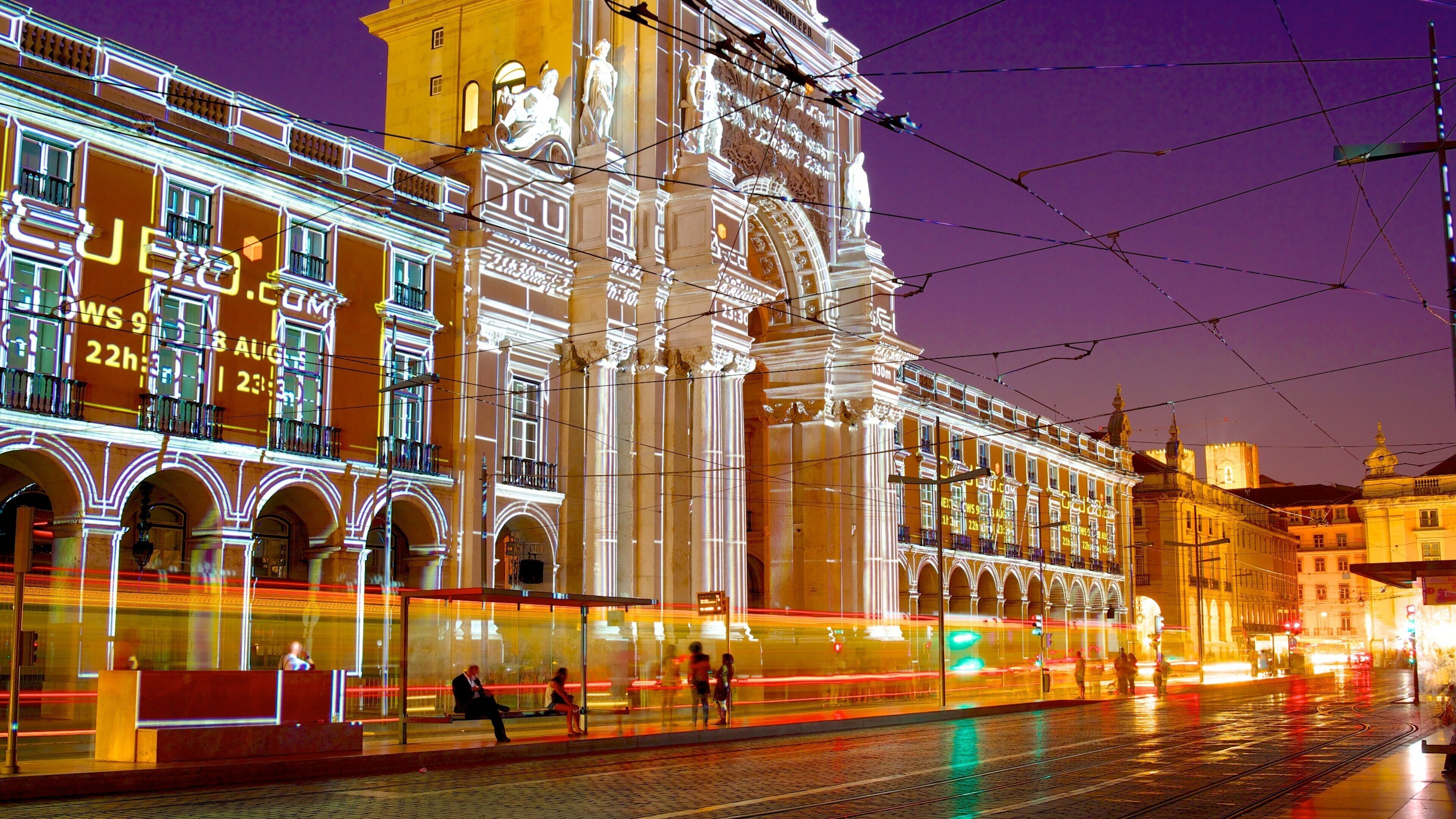 A brightly lit historic building with classical architecture, tram tracks, and a busy street scene at night.