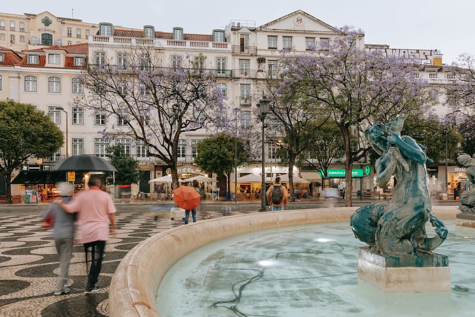 A fountain with statues, surrounded by a tiled plaza with buildings in the background.