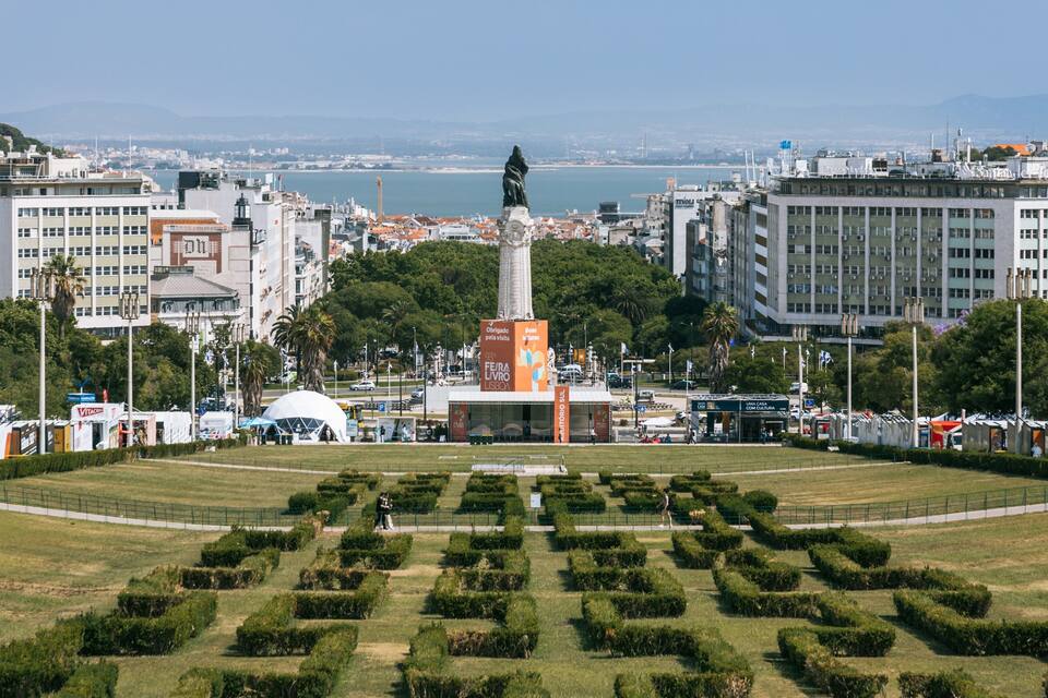 Un paisaje urbano con un monumento central, un parque amplio y abierto con setos podados, y una masa de agua al fondo.