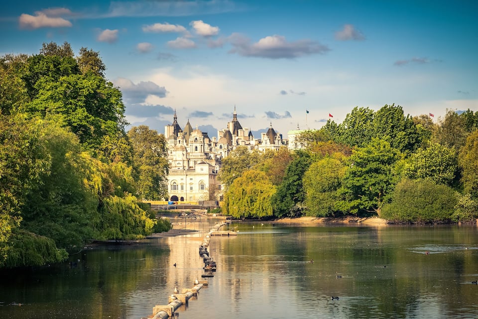 A river with a stone bridge, lush greenery, and a historic building with multiple spires in the background.