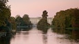 A calm lake with ducks, surrounded by trees and a large building in the background.