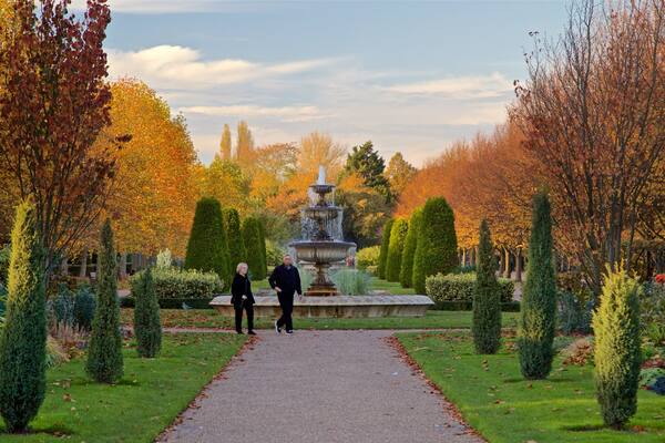 Ein Garten mit einem zentralen Brunnen, umgeben von ordentlich geschnittenen Hecken und Bäumen mit Herbstlaub.