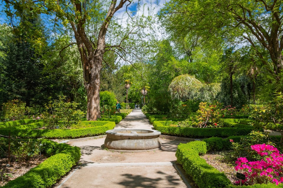 Ein Garten mit einem zentralen Kiesweg, gesäumt von ordentlich geschnittenen Hecken und verschiedenen blühenden Pflanzen.