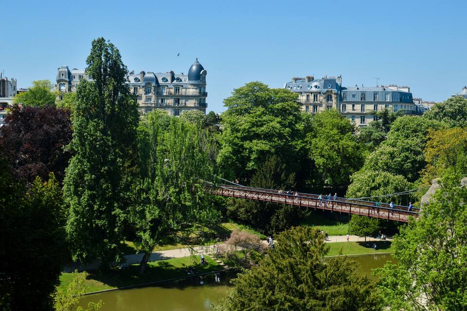 Um parque com uma ponte suspensa, vegetação exuberante e edifícios históricos ao fundo.