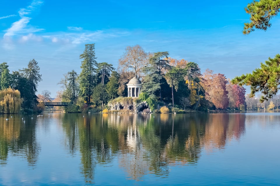 A tranquil lake with a gazebo, autumn trees, and a clear blue sky.