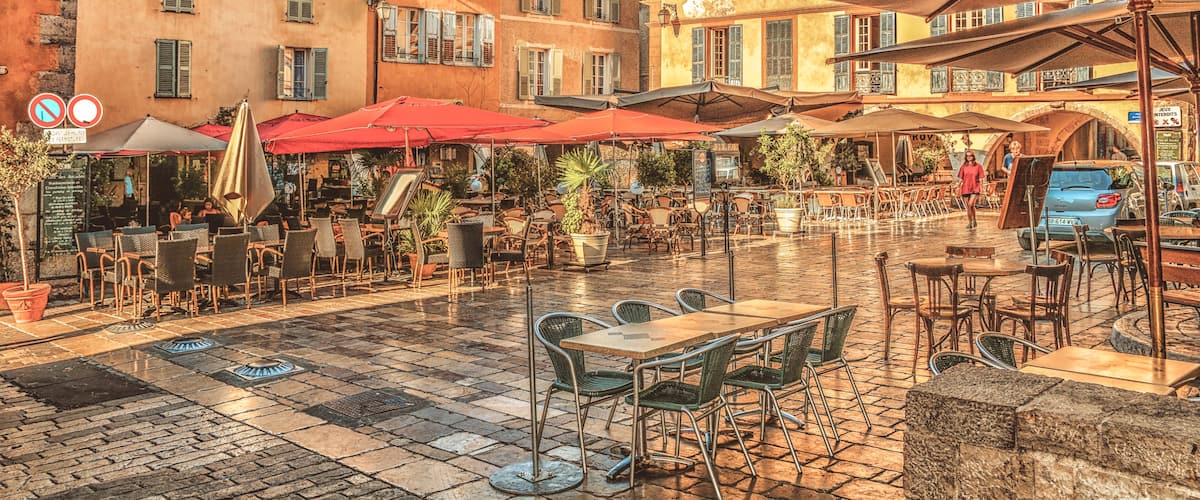 An outdoor café area with tables and chairs, red umbrellas, and buildings with balconies.