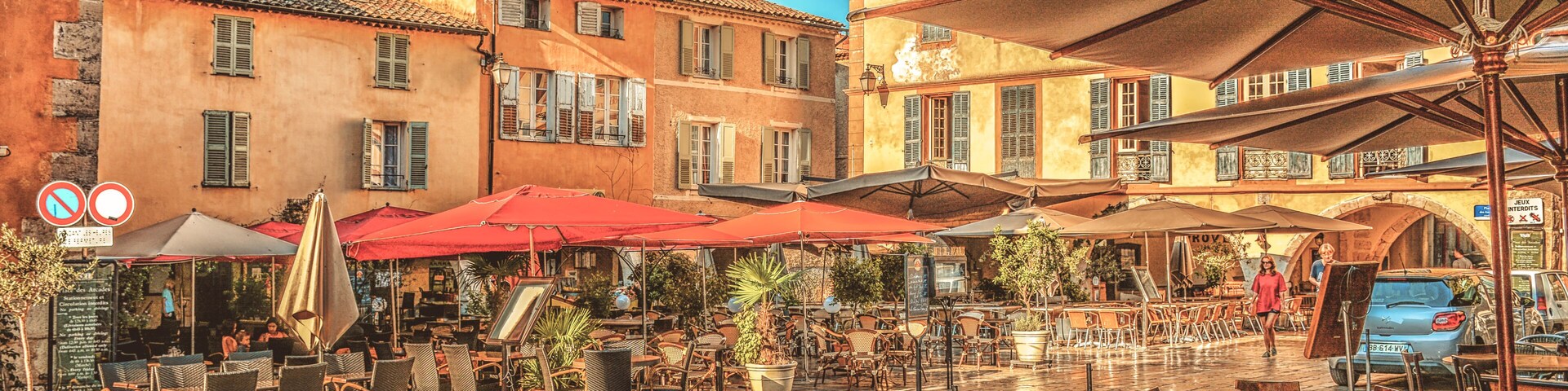 An outdoor café area with tables and chairs, red umbrellas, and buildings with balconies.