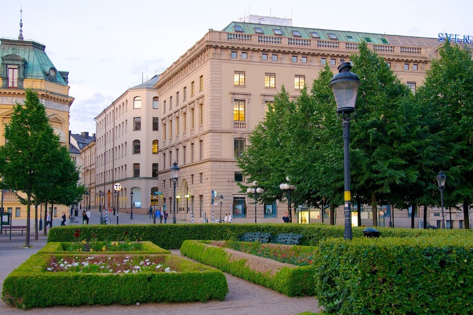Un jardín formal con setos perfectamente recortados y una gran variedad de flores. Al fondo, se aprecia un edificio histórico de grandes dimensiones con numerosas ventanas y tejado verde.