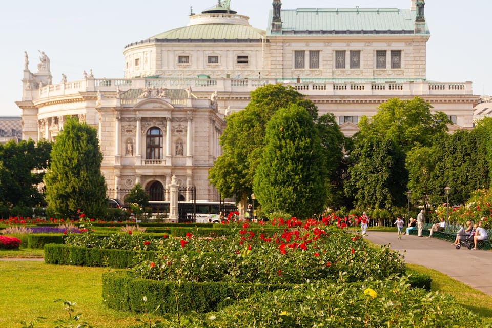 A grand building with a dome and statues, surrounded by well-manicured gardens and trees.