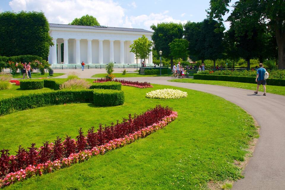 Un jardín cuidado con césped impecable, parterres de flores y un edificio clásico con columnas.