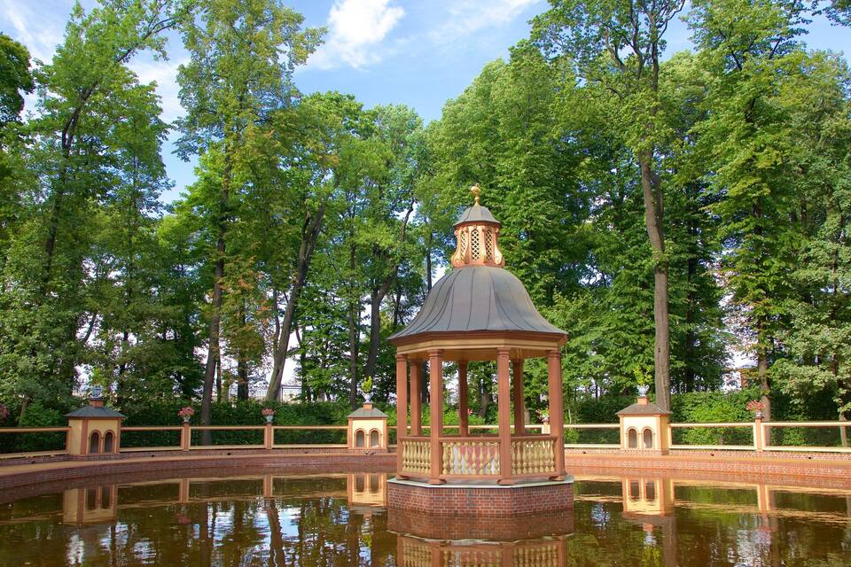 A gazebo with a dome roof surrounded by a brick pond, set in a park with tall trees and a clear sky.