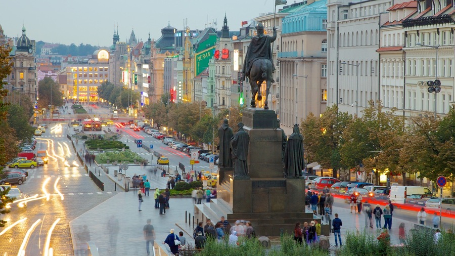 En travl gade i byen med en statue af en ridende mand, omgivet af historiske bygninger og en sporvognslinje.
