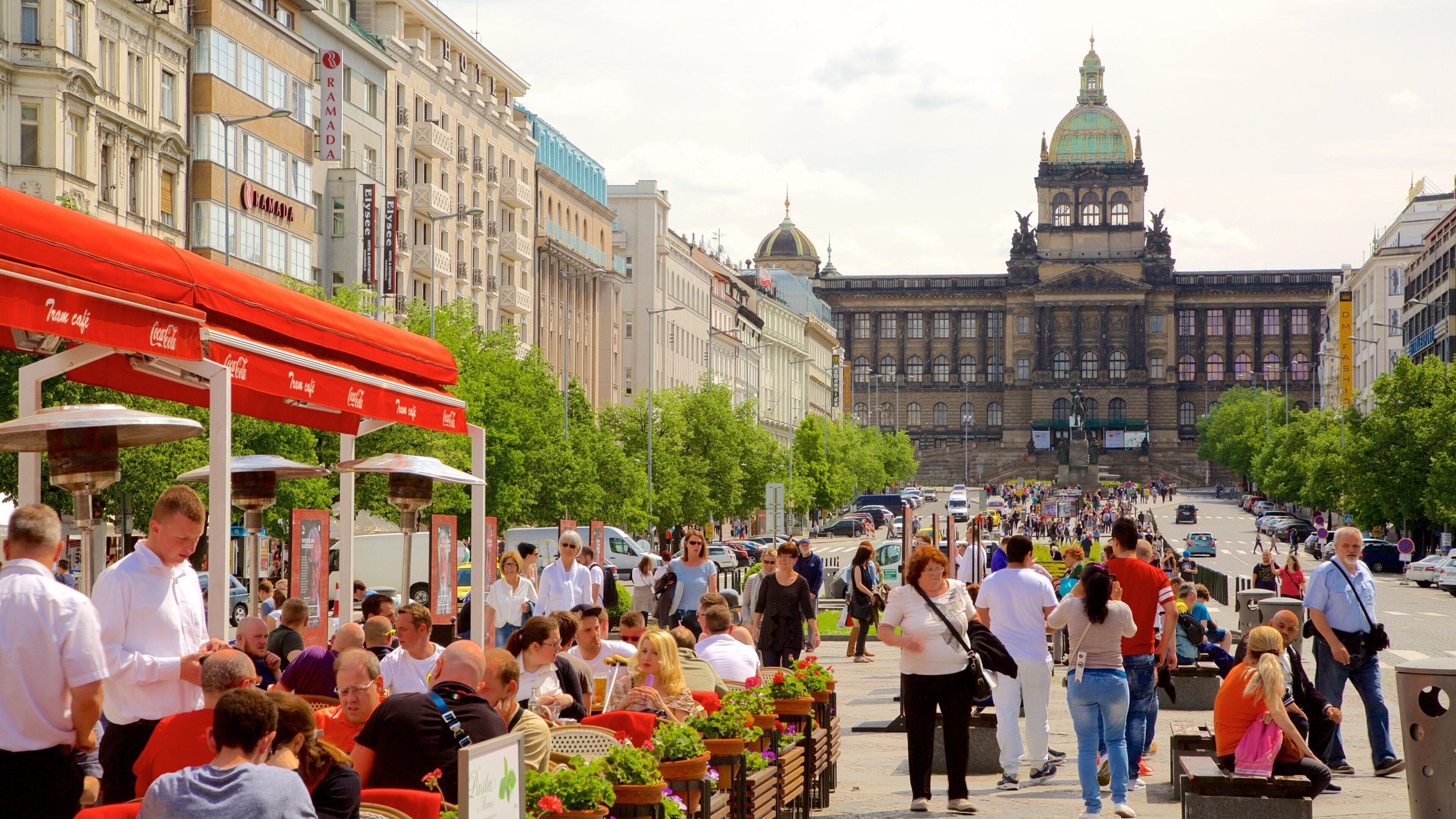 Una calle concurrida, gente, cafés al aire libre y un gran edificio histórico al fondo.
