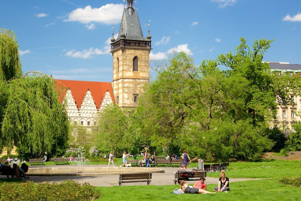 A park with a historic building featuring a tower and spires, surrounded by lush greenery and people enjoying the outdoors.