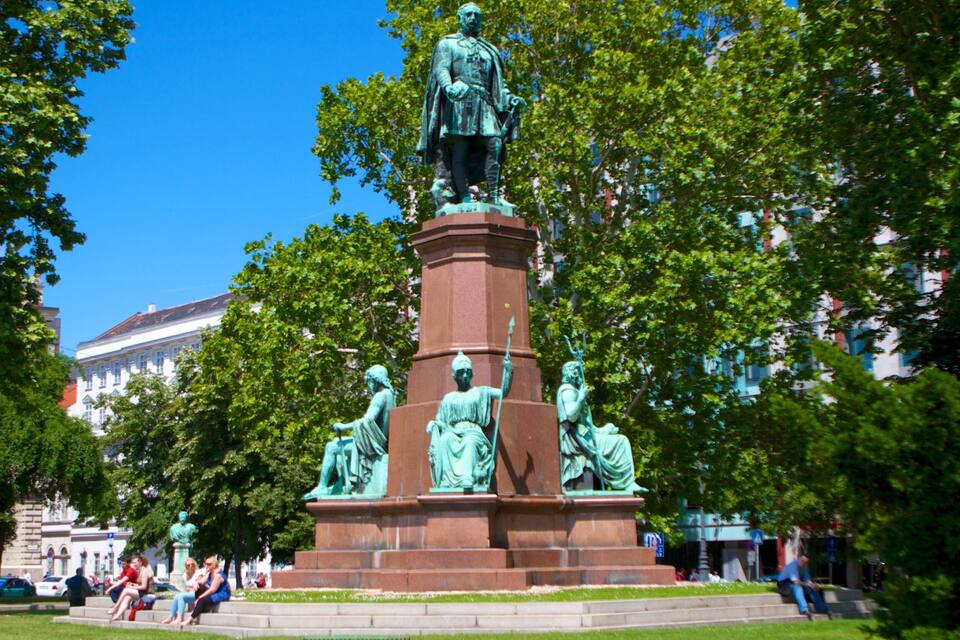 Una estatua de bronce de una figura histórica sobre un pedestal, con cuatro estatuas más pequeñas a su base, rodeada de árboles y edificios al fondo.