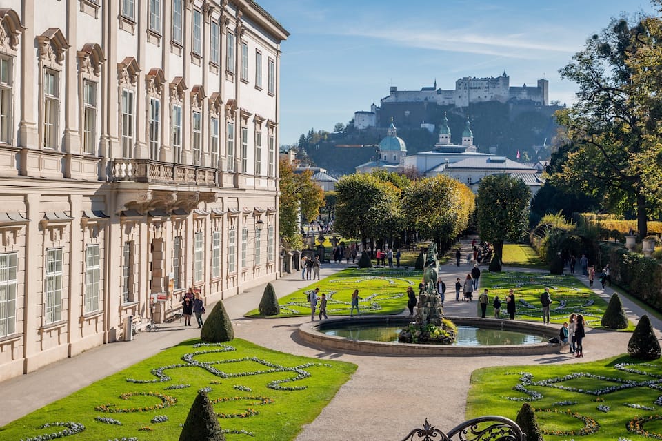 A historic building with ornate architecture, a well-maintained garden with geometric flower beds, and a castle in the background.