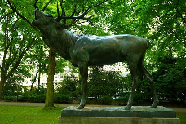 Eine Bronzestatue eines Wolfes in einem Park mit Bäumen und einem Gebäude im Hintergrund.