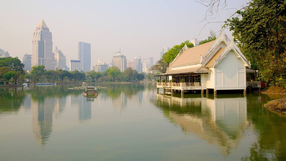 Um lago tranquilo com uma casa tradicional sobre palafitas, rodeado por vegetação exuberante e um horizonte urbano ao fundo.