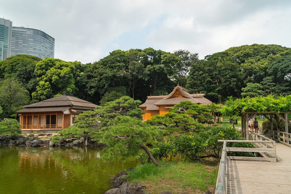 A traditional Japanese garden with a wooden bridge, a pond, and buildings with thatched roofs.