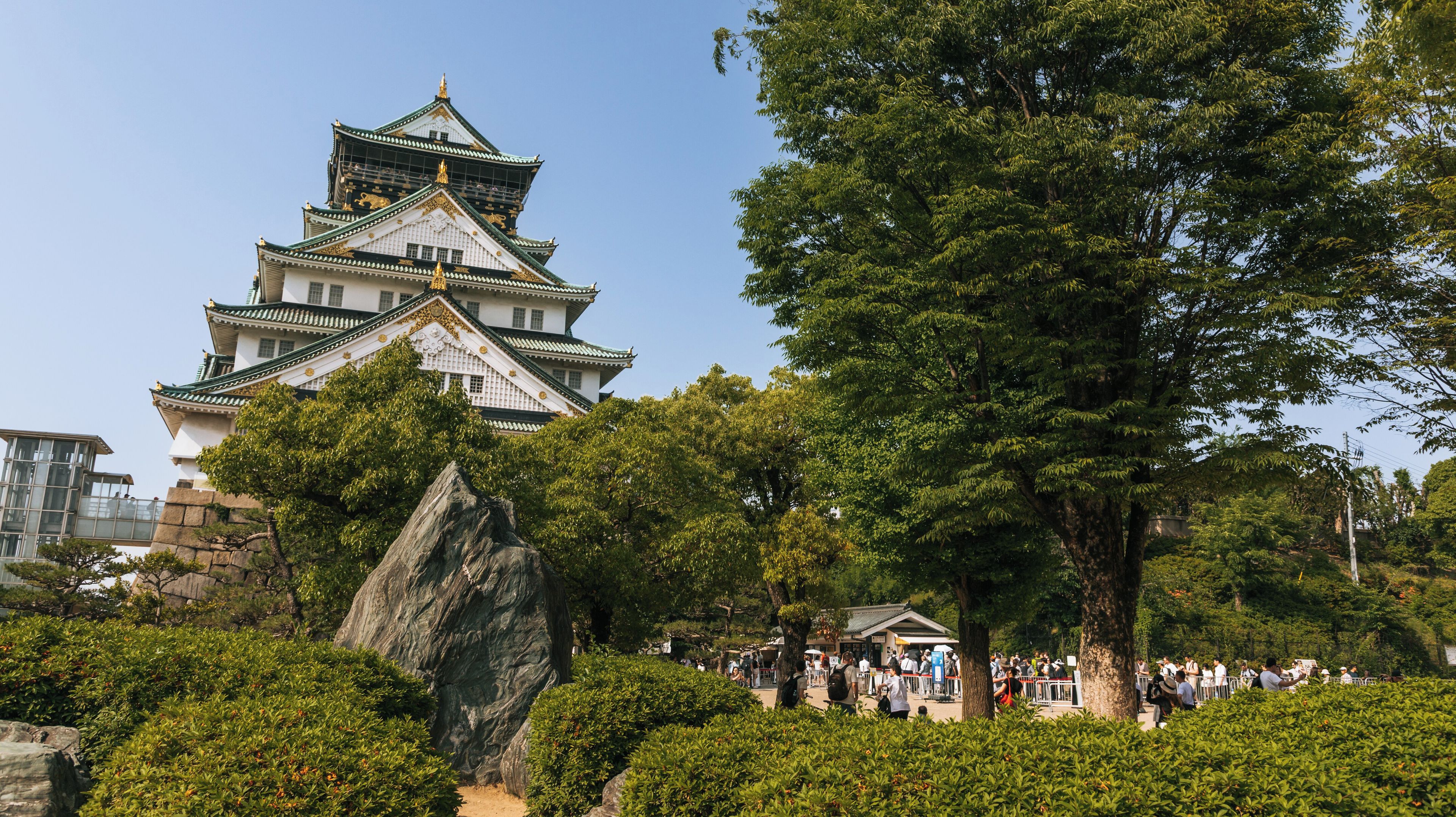 Un castell japonès tradicional amb un teulat de diversos nivells, envoltat de vegetació exuberant i una gran formació rocosa.