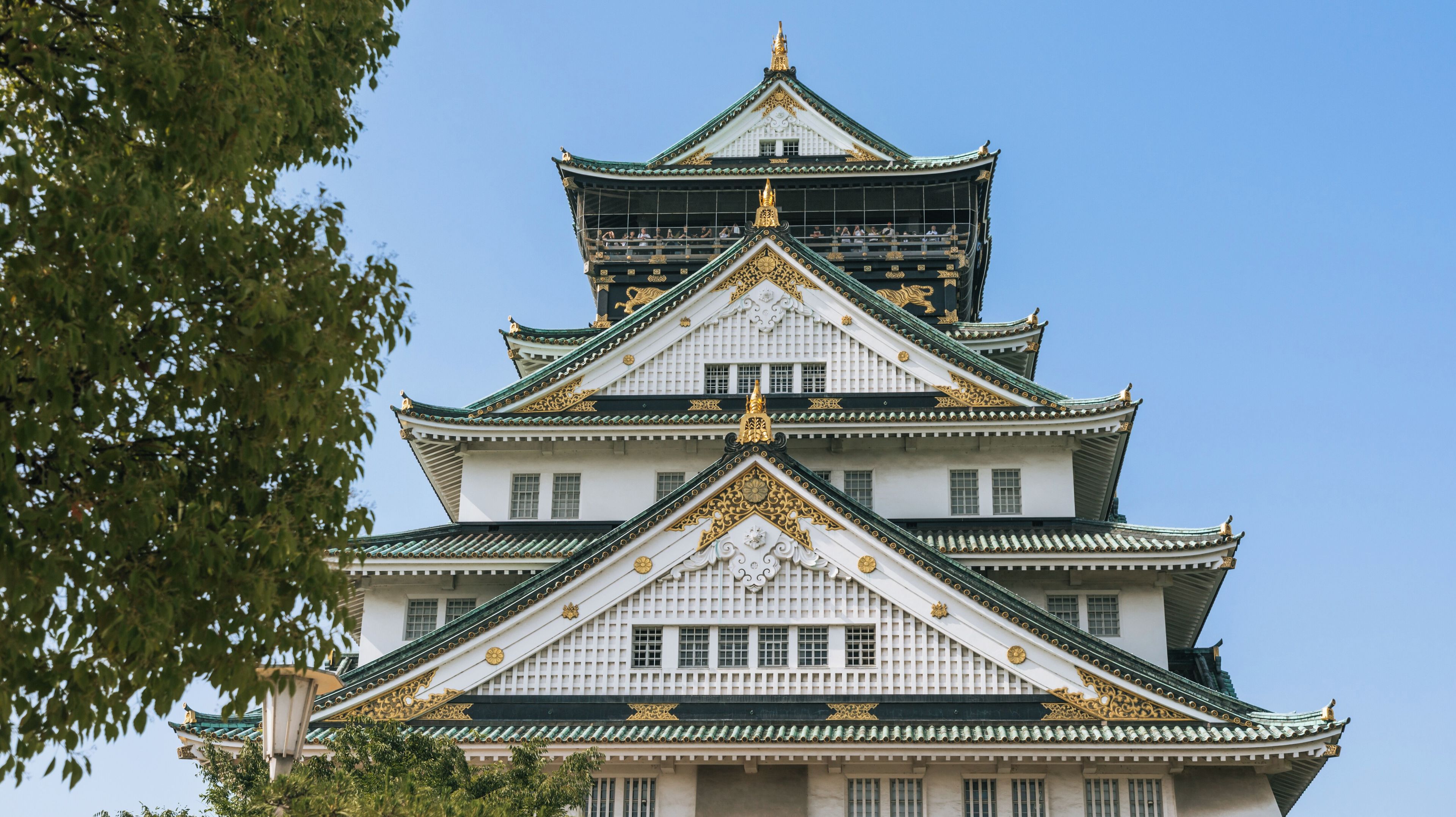 Un castell japonès tradicional de diverses plantes, amb detalls daurats i un cel blau clar al fons.