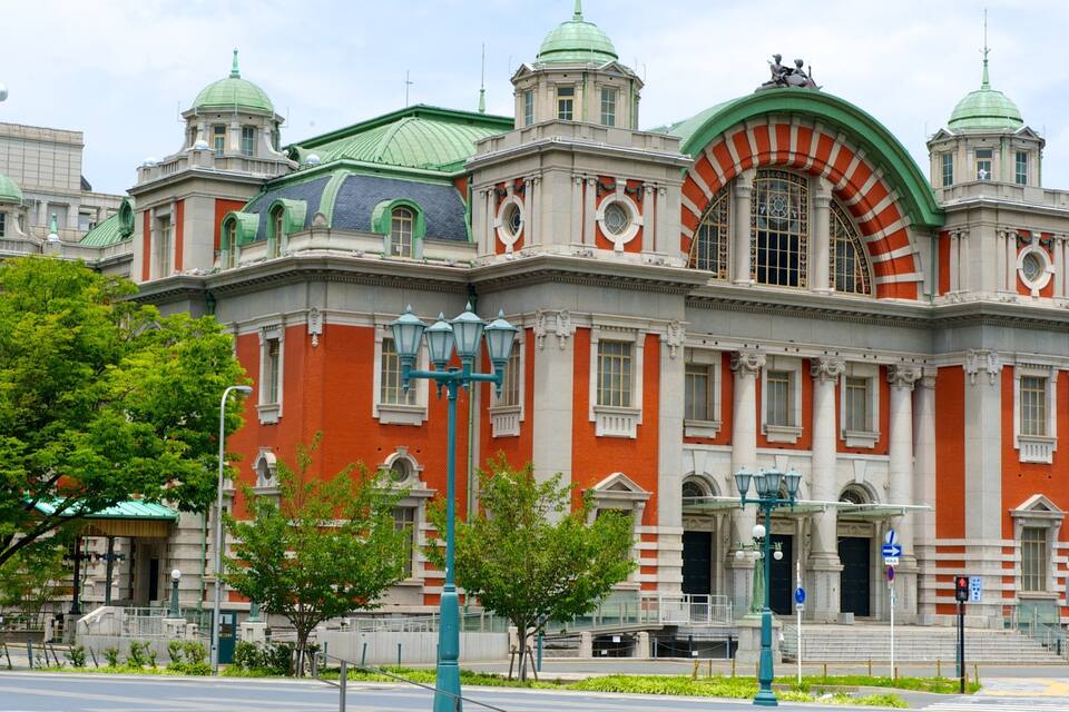 A historic building with red brick and green domes, surrounded by trees and modern buildings.