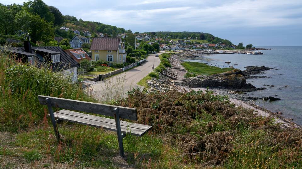 A coastal town with a bench overlooking the sea, houses along a road, and rocky shoreline.