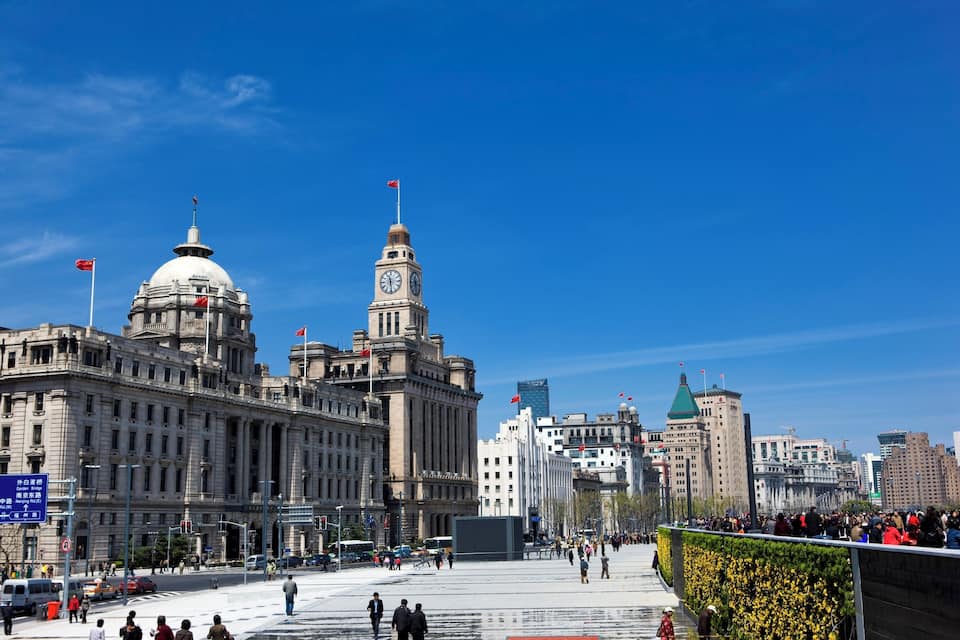 Un edificio grande y antiguo con una torre de reloj y una cúpula, rodeado de modernos rascacielos y una plaza concurrida llena de gente.
