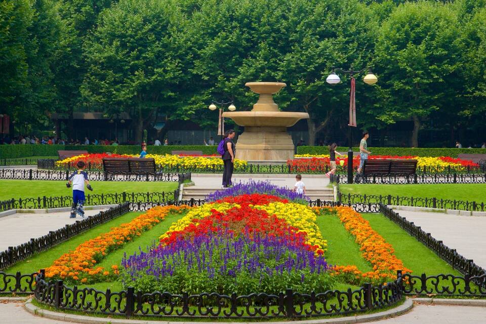A park with a central fountain, colorful flower beds, benches, and a black wrought iron fence.