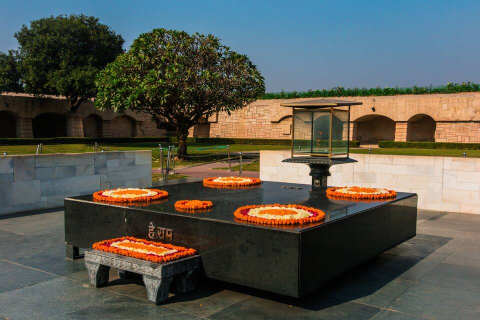 A memorial with a central black table surrounded by flower arrangements, set against a stone wall with a tree and a clear sky.