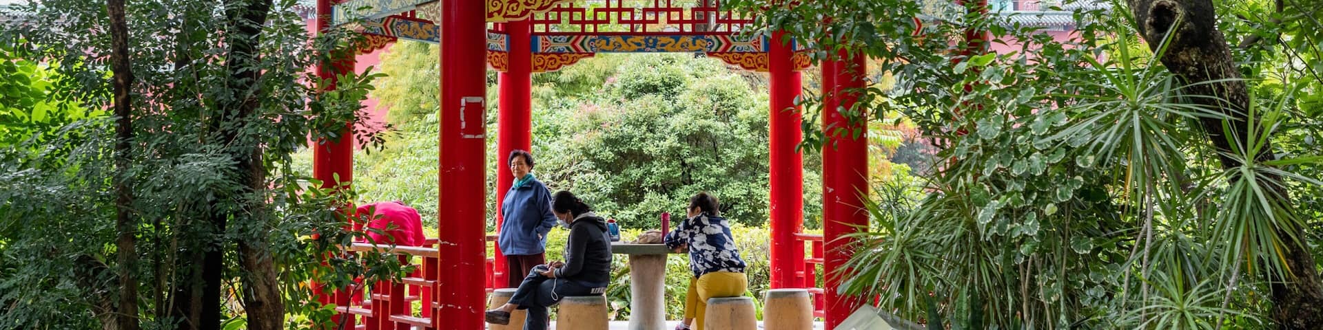 A traditional red and green pavilion with intricate designs, surrounded by lush greenery and trees.